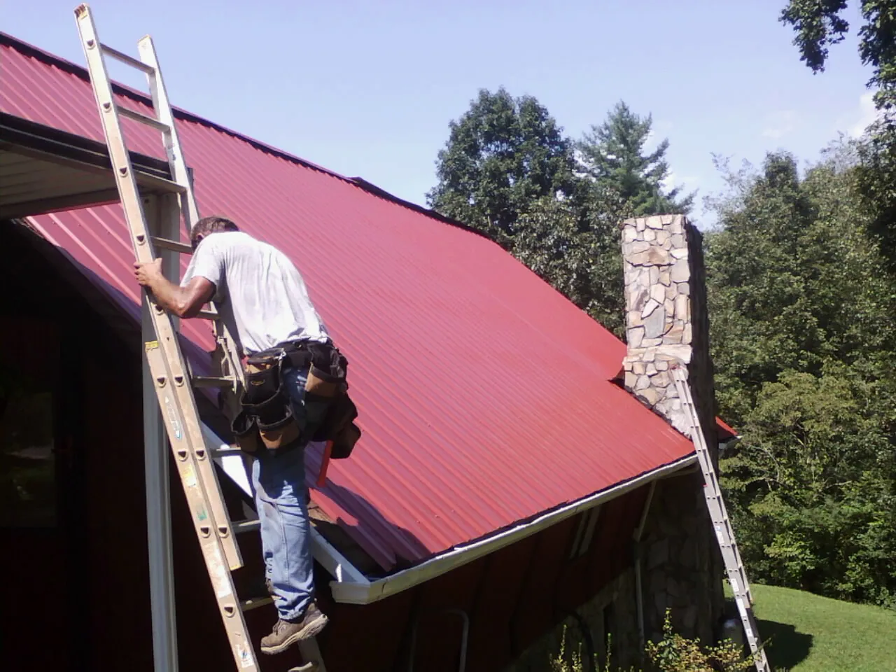 Roofer on a ladder beside a red metal roof and stone chimney — Soffit & Fascia Repair by Roofing Brothers in Bedrock